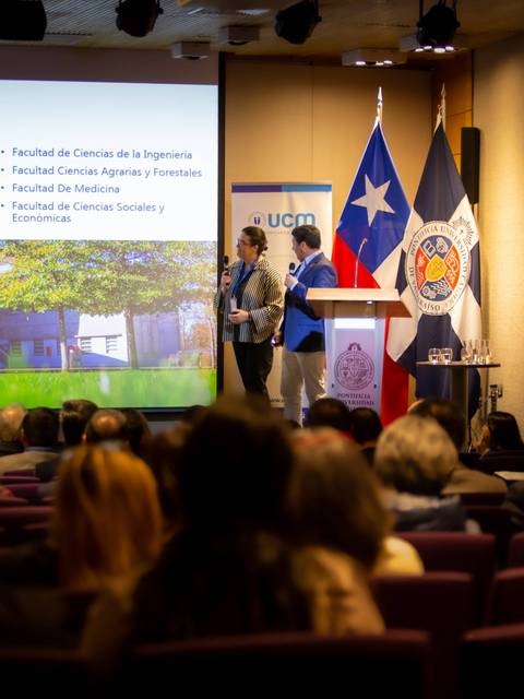 Una presentación en una sala con varias facultades expuestas en una pantalla y un público atento.