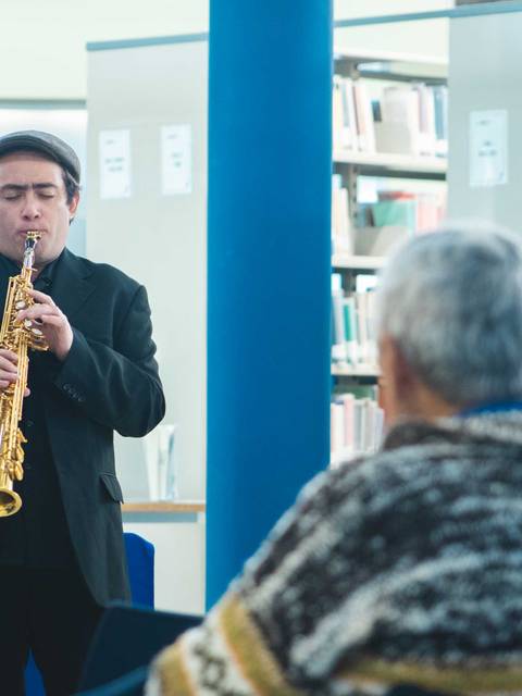 Un músico toca el saxofón en una biblioteca ante un grupo de personas que lo observan atentamente.