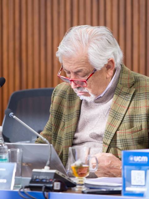 Un hombre mayor, con barba y gafas, revisa documentos en una mesa de trabajo en un entorno académico.