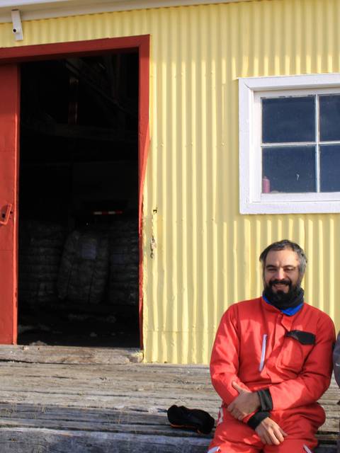 Dos hombres conversan frente a una casa amarilla con puerta roja.