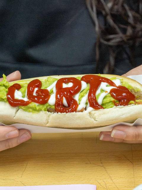 Un completo chileno decorado con palta y ketchup, sostenido por una persona.