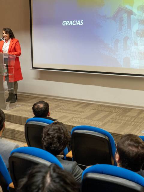 Una mujer en una chaqueta roja está presentando en un auditorio mientras una audiencia la escucha atentamente.