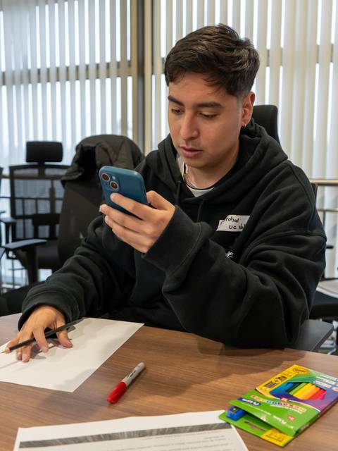 Un joven sentado en una mesa revisando su teléfono móvil mientras está rodeado de material de estudio.