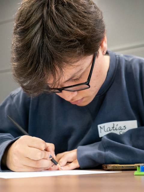 Un cabro joven, con lentes, está concentrado escribiendo en una hoja de papel.