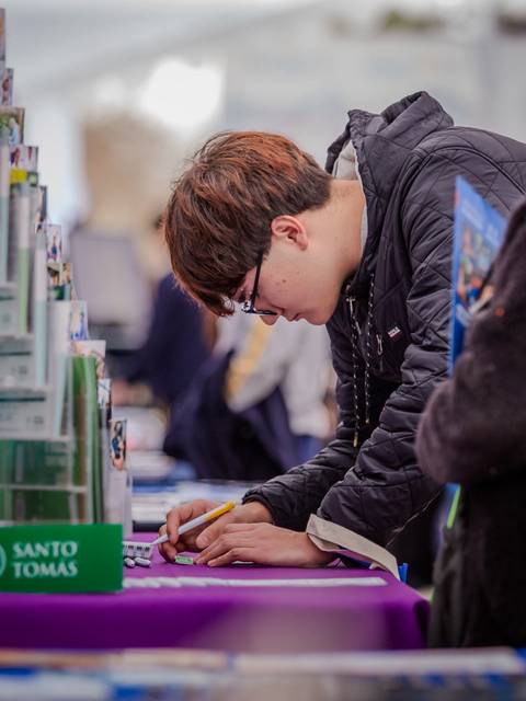 Un joven escribe concentrado en un stand mientras otras personas observan.