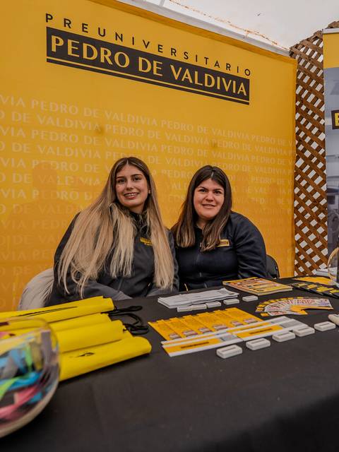 Dos chicas sonrientes en un stand promocional del preuniversitario Pedro de Valdivia con artículos informativos y coloridos.