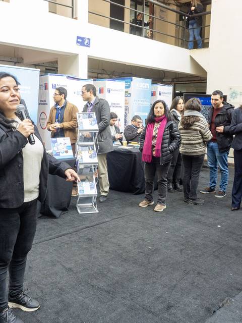 Una mujer habla al público en un evento con stands de presentación en un ambiente interior.