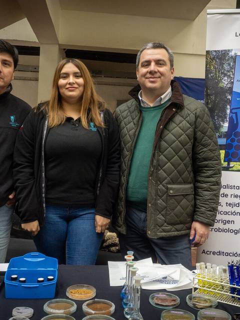Tres personas posan junto a un stand de laboratorio en un evento agrícola.