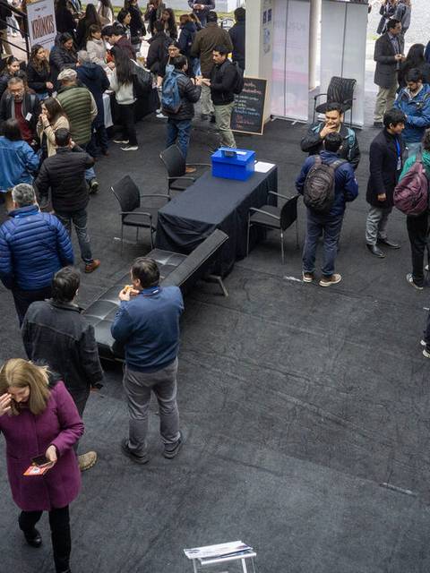 Un grupo grande de personas se congrega en un evento con stands informativos y mesas en un ambiente interior.