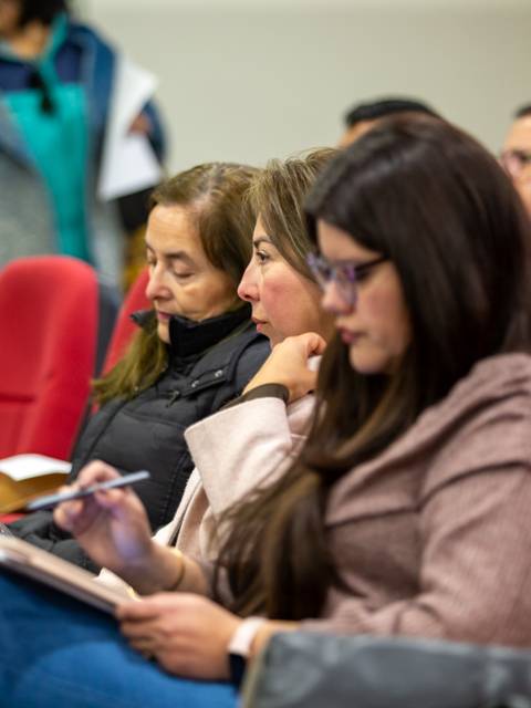 Un grupo de personas atentas escuchando en un evento, sentados en sillas rojas.