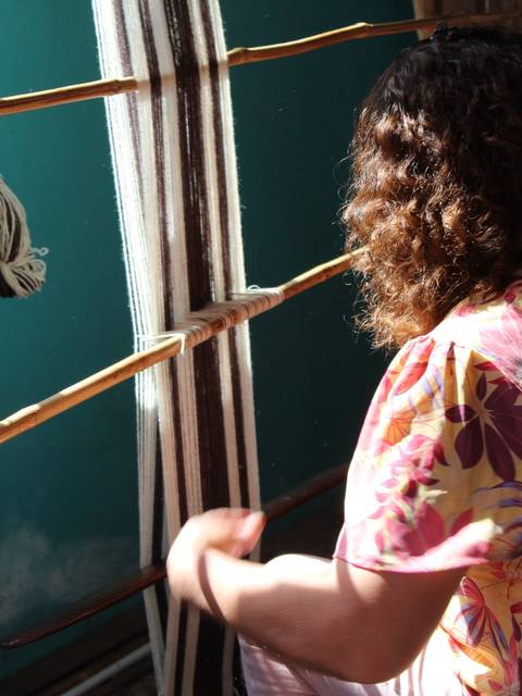 Una mujer tejiendo con hilos en un telar antiguo en una habitación iluminada por la luz del sol.