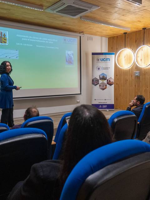 Una expositora presenta un tema ante un público atento en una sala moderna.