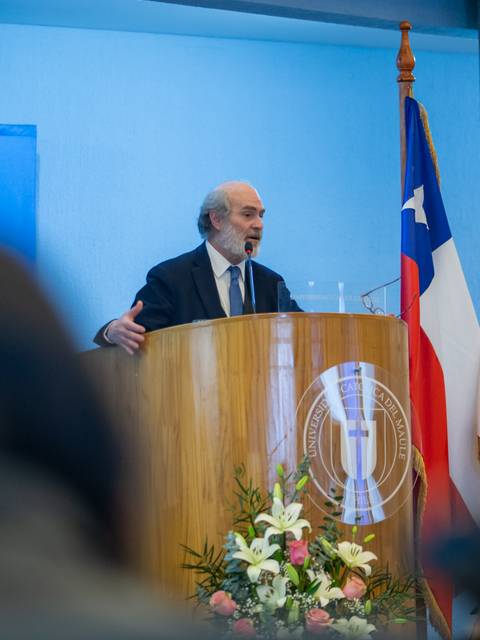 Un profesor da una charla en una ceremonia en la Universidad Católica de Chile, con banderas de fondo.