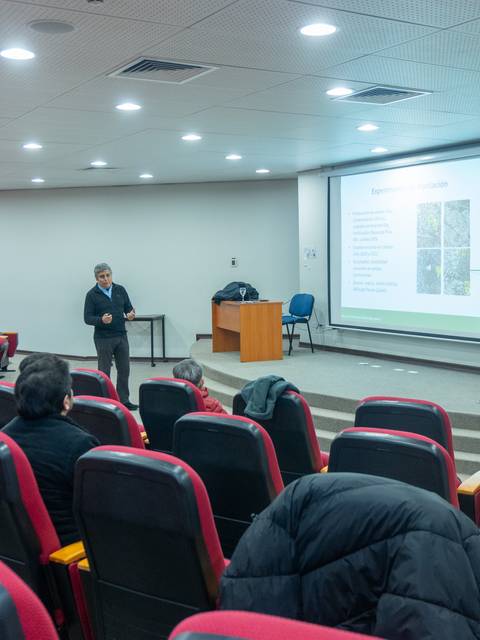 Un profesor está exponiendo en una sala con asientos rojos.