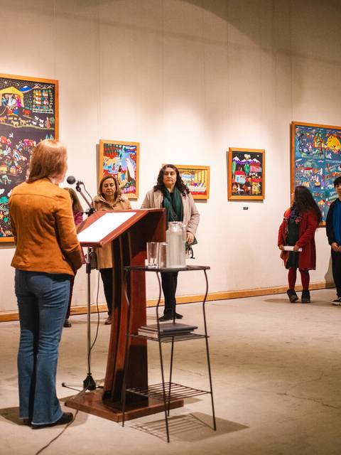 En una galería de arte, una mujer habla frente a un grupo de personas que observan pinturas coloridas en las paredes.