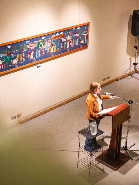 Una mujer presenta en un escenario mientras se exhiben pinturas en la pared.