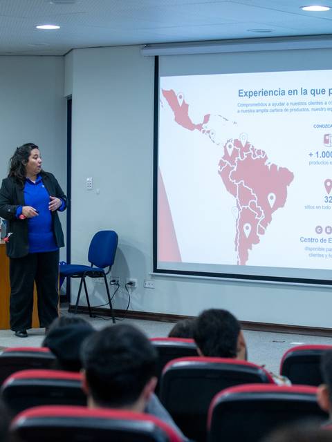 Una mujer presenta información sobre la experiencia de una empresa en Latinoamérica en una sala llena de estudiantes.
