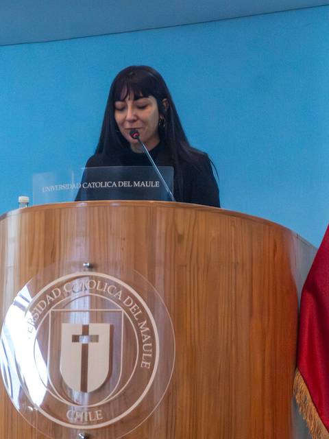 Una mujer está hablando en un podio con el logo de una universidad chilena y una bandera de Chile al lado.