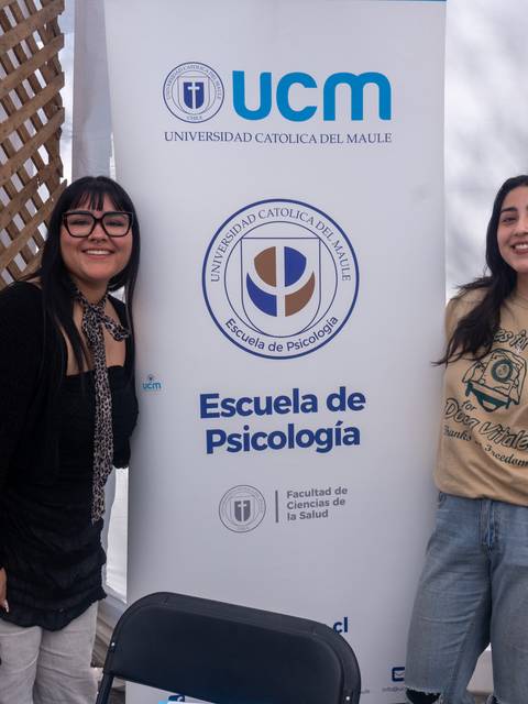 Dos mujeres sonrientes posan junto a un banner de la Universidad Cátolica del Maule y la Escuela de Psicología.