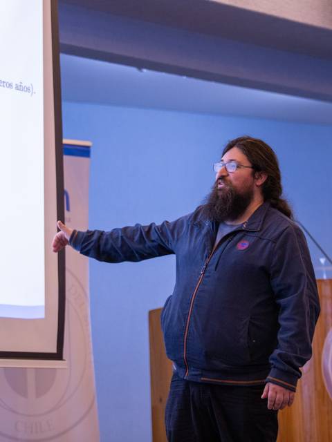 En la imagen se observa a un hombre presentando una charla en un auditorio con banderas chilenas de fondo.