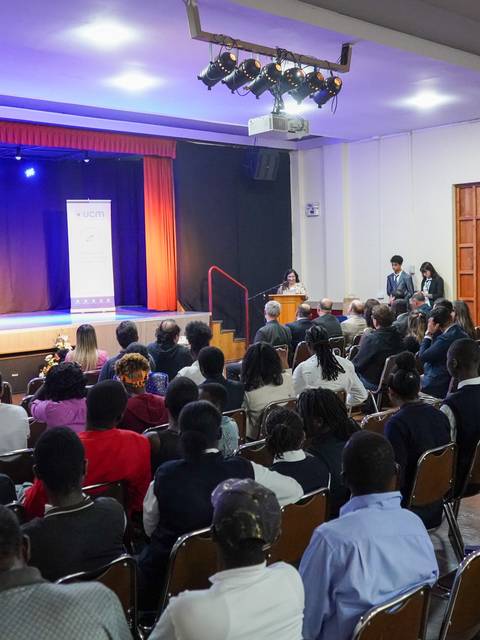 Una conferencia en un lugar amplio con un público atento enfrente de un escenario iluminado.