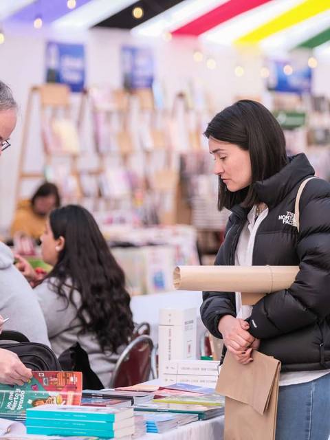 En una feria del libro, un hombre y una mujer conversan sobre libros mientras otros lectores exploran.