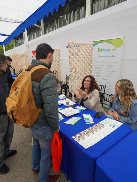 En una feria, un grupo de personas interactúa en un stand, donde dos mujeres presentan información a un potencial interesado.
