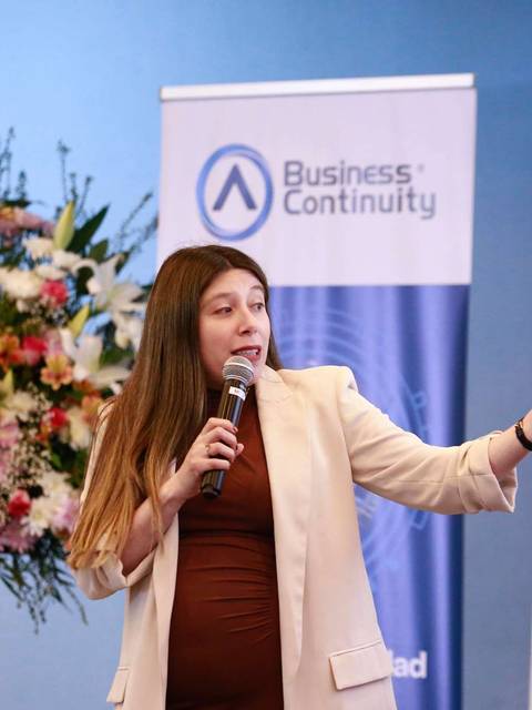 Una mujer presenta en un evento mientras sostiene un micrófono y señala hacia un lado.