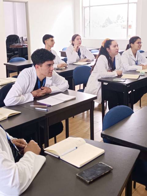 Un grupo de estudiantes con batas blancas prestando atención en una sala de clases.
