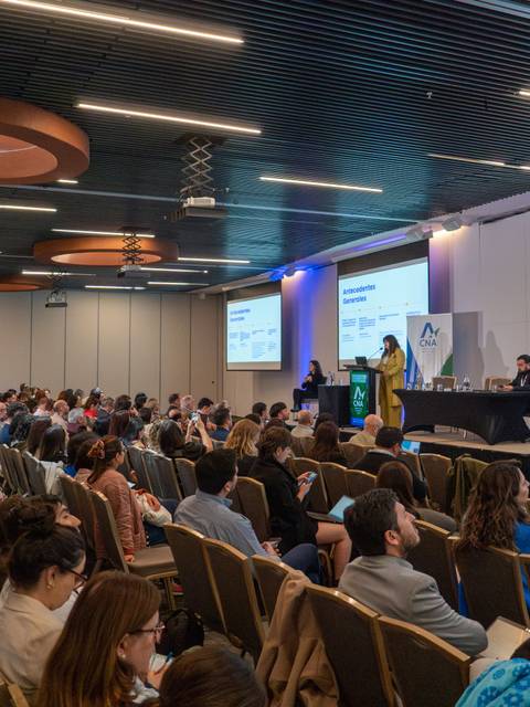 Una conferencia llena de gente escuchando a una expositora en el escenario.