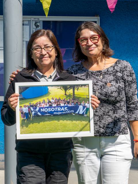Dos mujeres posan sonrientes sosteniendo un marco con una foto de grupo en un ambiente colorido.