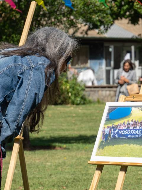 Una mujer observa atentamente una foto en un marco, mientras en el fondo hay otras personas disfrutando del día al aire libre.