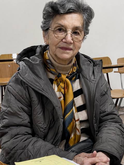 Una señora mayor con bufanda y gafas sonríe mientras está sentada en una sala vacía.
