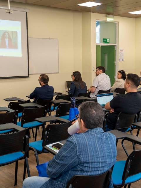 Una presentación en un aula con varios asistentes, donde una mujer expone frente a una pantalla.