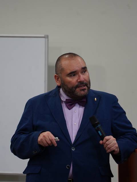 Un hombre vestido formalmente con corbata, presentando un tema en una sala de conferencias.