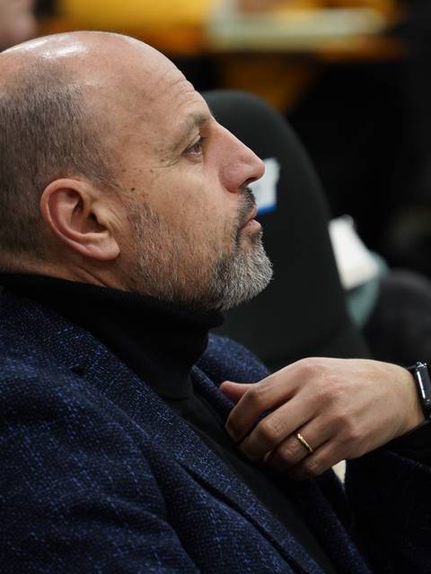 Un hombre con barba y chaqueta sentado, observando atentamente en una reunión.