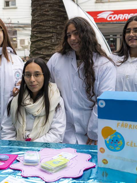 Un grupo de estudiantes vestidas con delantales blancos presentan una actividad educativa sobre células en una feria.