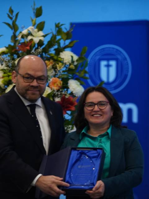 En una ceremonia, dos personas posan sonrientes, una de ellas sostiene un premio frente a un fondo decorado con flores.