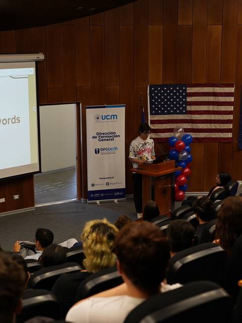 Un grupo de estudiantes asiste a una presentación en un auditorio con banderas de Estados Unidos y Australia de fondo.