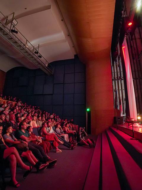 Un público entusiasta disfruta de una presentación en un teatro iluminado con luces rojas.
