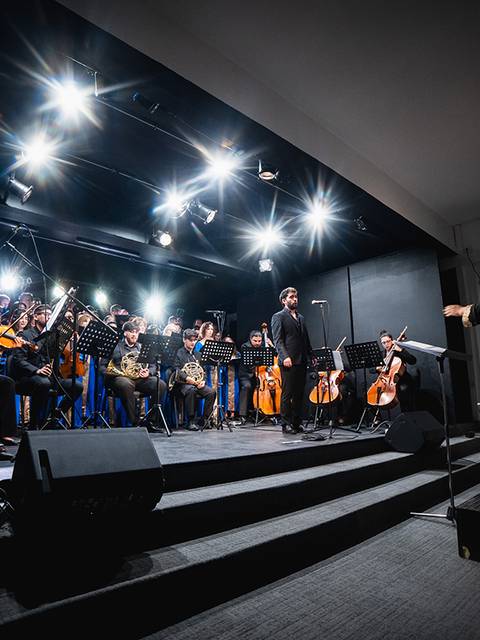 Orquesta tocando en un escenario iluminado con un director dirigiendo la música.