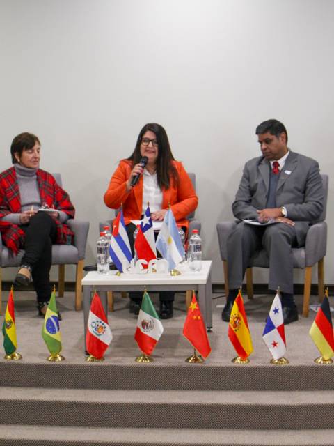 Un panel de discusión con cinco personas vestidas formalmente, sentados en un escenario con banderas de varios países.