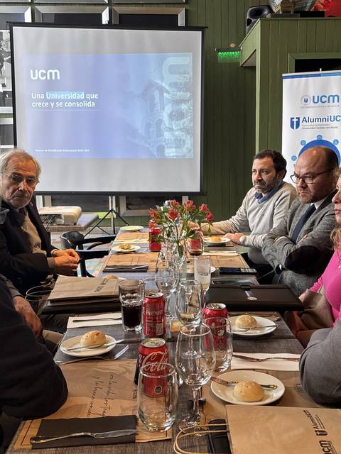 Un grupo de personas en una reunión en torno a una mesa con comida y un proyector mostrando la presentación de la UCM.