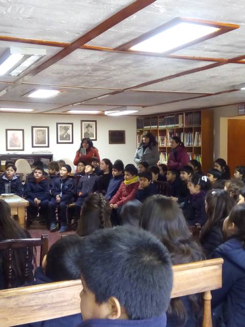 En una biblioteca, un grupo de niños escucha a un presentador frente a una mesa.