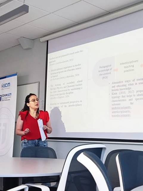 Una joven presentando en una sala de clases con proyector y sillas en un ambiente académico.
