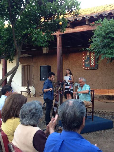 Un evento comunitario al aire libre donde varias personas están sentadas y observando a dos personas en el escenario.