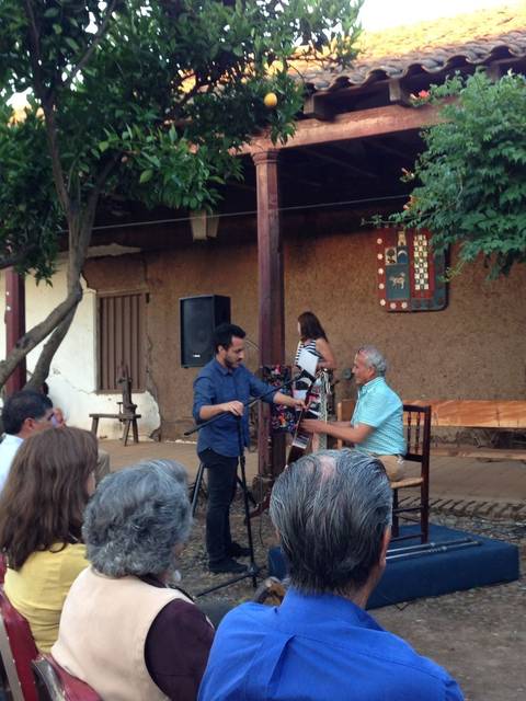 Un evento cultural al aire libre con un público atento en un patio con árboles y decoración rústica.