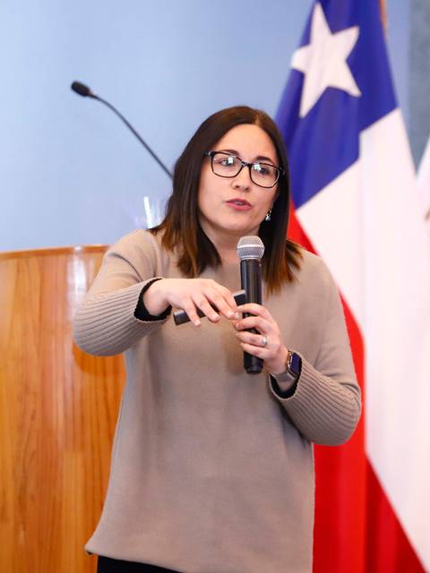 Una mujer presenta un tema con entusiasmo mientras sostiene un micrófono frente a una audiencia.