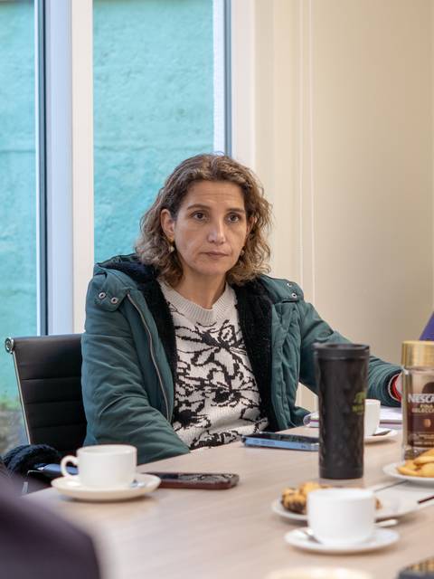 Una mujer sentada en una reunión, mirando atentamente mientras otros conversan.