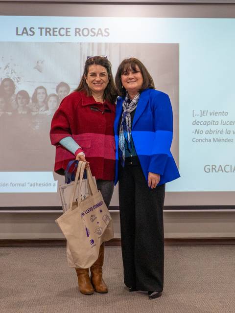 Dos mujeres posando sonrientes en un evento, con un fondo que muestra la frase 'Las Trece Rosas'.